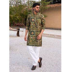 A man wearing a green and gold patterned kurta with white trousers and dark loafers, standing outdoors on a light gravel path.