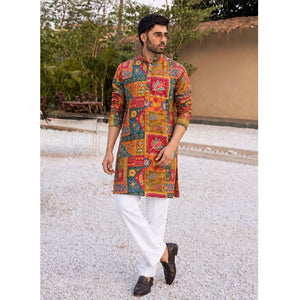 A man wearing a vibrant, multicolored patchwork-print kurta with white trousers and black loafers, standing outdoors on a light gravel path with a house and greenery in the background.