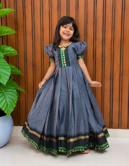 A young girl modeling a dark gray-blue traditional Indian dress with green and gold embroidery, standing in front of a brown wooden slatted wall.