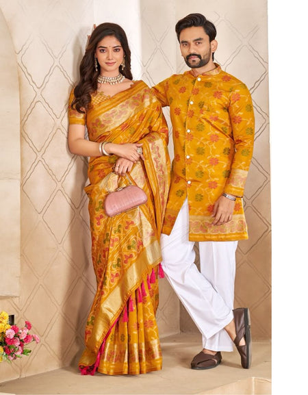 A man and a woman posing in traditional Indian attire. The woman wears a yellow and gold Banarasi silk saree with pink and orange floral patterns and a pink clutch.