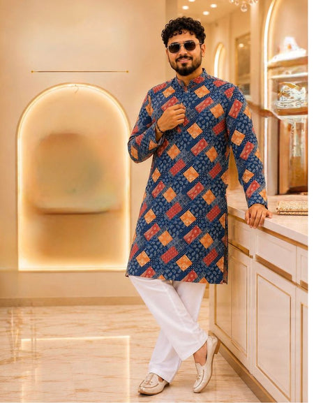 A man wearing a blue patterned kurta with red and orange diamond motifs, paired with white trousers and beige loafers, standing in a modern, warmly lit boutique.
