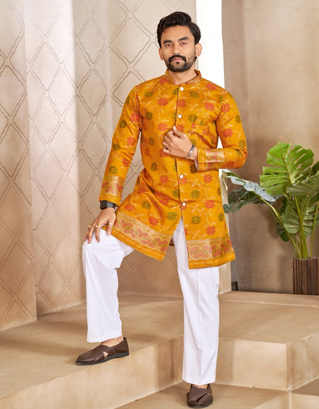 A man modeling a bright yellow-orange kurta with intricate multicolor floral patterns and white trousers, standing on beige stone steps in front of a patterned beige wall.