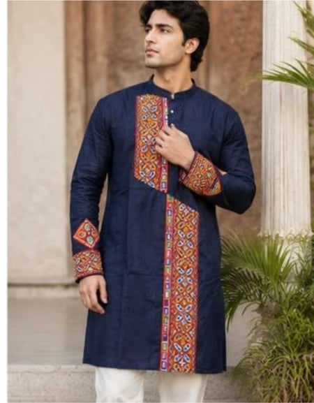 A man wearing a navy blue kurta with colorful geometric embroidered panels on the front and cuffs, paired with white trousers, standing outdoors in front of a stone column and plants.