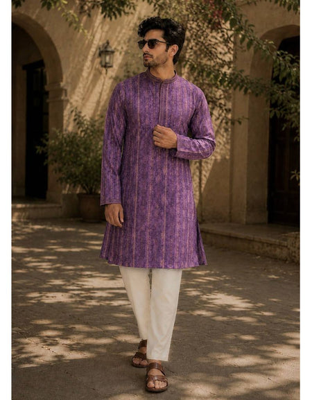 A man wearing a festive wear ethnic traditional Indian purple patterned kurta with white trousers and brown sandals, standing outdoors in front of a beige stone building with arches and leafy branches.
