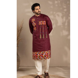 A man modeling a festive wear Navratri deep maroon kurta with colorful geometric embroidery on the chest and hem, paired with white trousers and brown sandals, set against a warm beige background with soft leaf shadows.