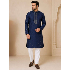 A man modeling a navy blue kurta with small gold motifs and a patterned mandarin collar, paired with white trousers and brown shoes, set against a beige architectural background.
