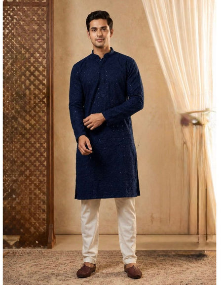 A man modeling a dark navy blue kurta with subtle embroidered patterns, paired with off-white trousers and brown shoes, standing in a warm-toned indoor setting with curtains.