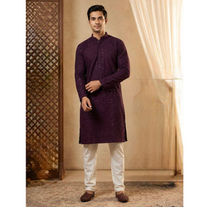 A man modeling a traditional Indian wine long-sleeve kurta with subtle embroidered patterns, paired with off-white trousers and brown shoes, standing in a warm-toned indoor setting with a patterned screen and sheer curtain.