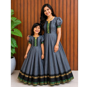 A woman and a young girl wearing matching dark gray traditional Anarkali-style dresses with black, green, and gold border details, standing in front of a wooden paneled wall.