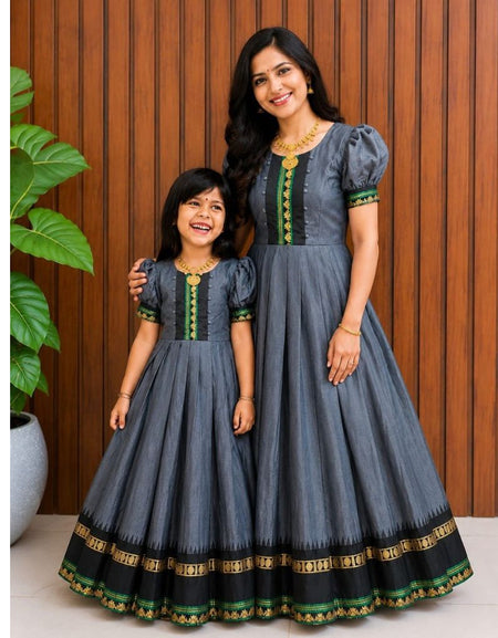 A woman and a young girl wearing matching dark gray traditional Anarkali-style dresses with black, green, and gold border details, standing in front of a wooden paneled wall.