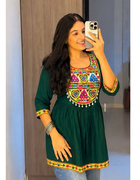 A woman taking a mirror selfie wearing a dark green cotton kurta with colorful embroidered patterns around the neckline and sleeve cuffs, paired with blue jeans.