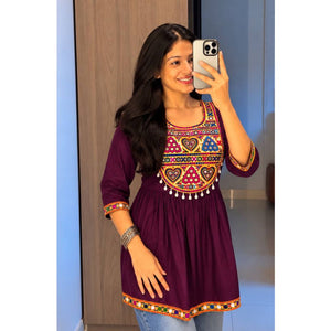 A woman taking a mirror selfie wearing a deep purple kurta with colorful geometric and heart embroidery around the neckline and sleeve cuffs, paired with light blue jeans.