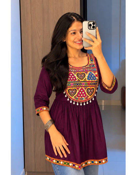 A woman taking a mirror selfie wearing a deep purple kurta with colorful geometric and heart embroidery around the neckline and sleeve cuffs, paired with light blue jeans.
