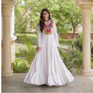 A woman wearing a long white maxi dress with colorful embroidered detailing on the chest stands in a garden corridor framed by beige pillars.
