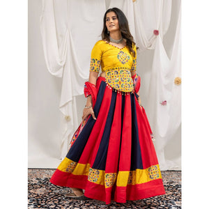 A woman models a traditional Indian ethnic outfit consisting of a bright yellow embroidered blouse and a voluminous red and navy blue skirt with yellow border details, set against a draped white fabric backdrop.