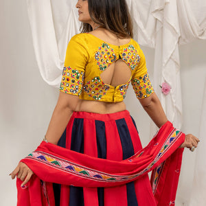 A woman modeling a traditional Indian outfit, shown from the back. She is wearing a bright yellow blouse with colorful floral embroidery and a back cutout, paired with a red and navy blue striped skirt and holding a red dupatta with a colorful border.