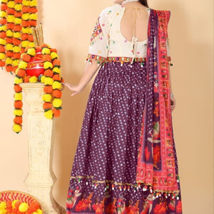A woman wearing a traditional Indian outfit with a purple patterned skirt, a white embroidered blouse, and a red patterned dupatta, standing in front of a beige wall with decorative garlands