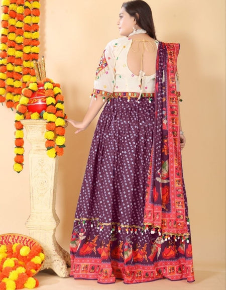 A woman wearing a traditional Indian outfit with a purple patterned skirt, a white embroidered blouse, and a red patterned dupatta, standing in front of a beige wall with decorative garlands