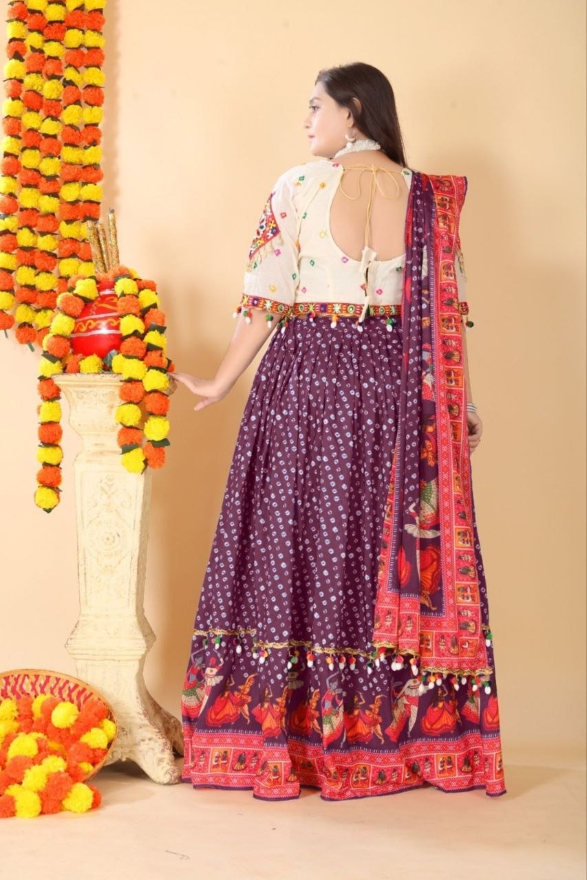 A woman wearing a traditional Indian outfit with a purple patterned skirt, a white embroidered blouse, and a red patterned dupatta, standing in front of a beige wall with decorative garlands