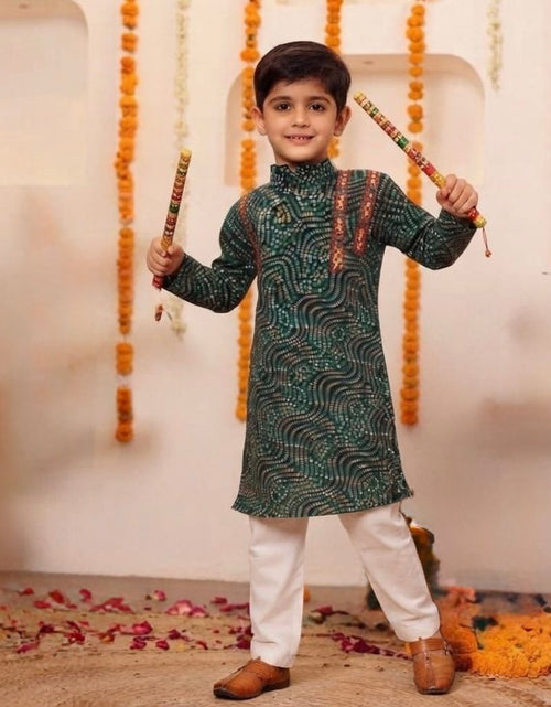 Load image into Gallery viewer, A young boy wearing a dark green patterned kurta and white trousers, holding traditional Indian sticks (dhaka) against a decorated indoor backdrop with marigold garlands.
