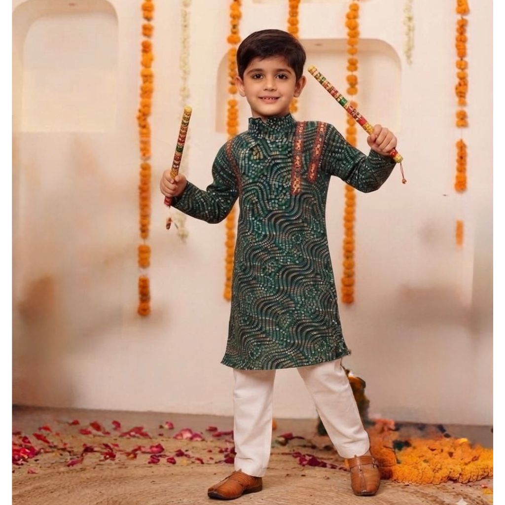 A young boy wearing a dark green patterned kurta and white trousers, holding traditional Indian sticks (dhaka) against a decorated indoor backdrop with marigold garlands.