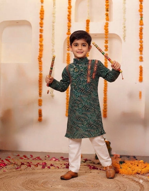 Load image into Gallery viewer, A young boy stands in a decorated indoor setting wearing a dark green patterned kurta with white pants and brown shoes, holding two colorful sticks. The background features a cream-colored wall with arched recesses, hanging orange marigold garlands, and scattered flower petals on the floor.
