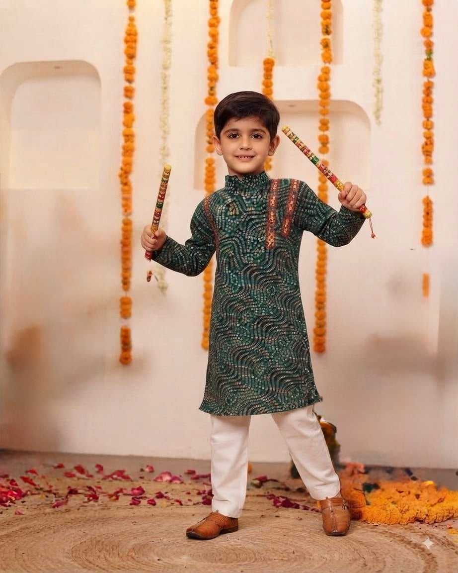 A young boy stands in a decorated indoor setting wearing a dark green patterned kurta with white pants and brown shoes, holding two colorful sticks. The background features a cream-colored wall with arched recesses, hanging orange marigold garlands, and scattered flower petals on the floor.