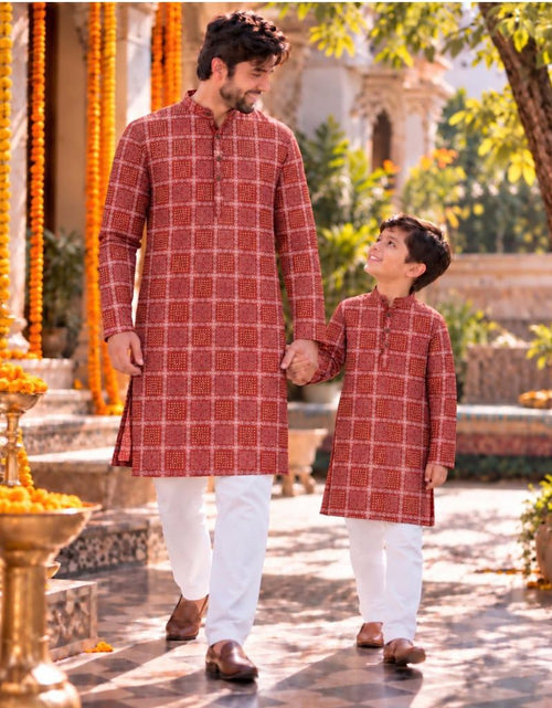 Load image into Gallery viewer, A man and a child walking hand in hand outdoors, wearing matching red patterned kurta sets with white pants. The background shows a sunlit courtyard with orange marigold garlands and architectural details.
