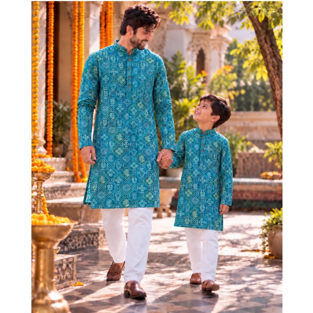 A man and a boy holding hands, wearing matching teal blue patterned kurta sets with white trousers, walking hand-in-hand in a traditional courtyard decorated with orange marigold garlands.
