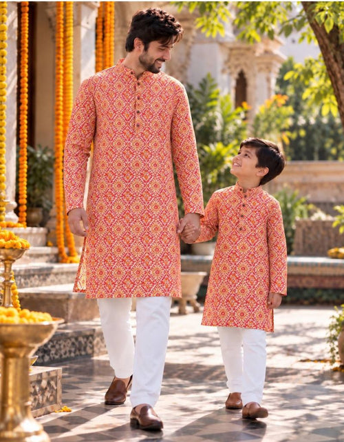 Load image into Gallery viewer, A man and a boy wearing matching peach-orange printed kurta sets and white trousers walking hand-in-hand in a garden courtyard decorated with yellow marigold garlands.
