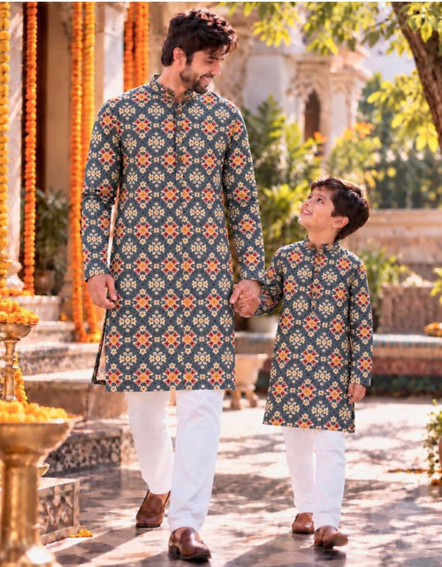 Load image into Gallery viewer, A man and a boy wearing matching printed kurta sets walk hand in hand in an outdoor courtyard. The long-sleeve shirts have a dark teal-blue base with a repeating orange and yellow geometric pattern, paired with white trousers.
