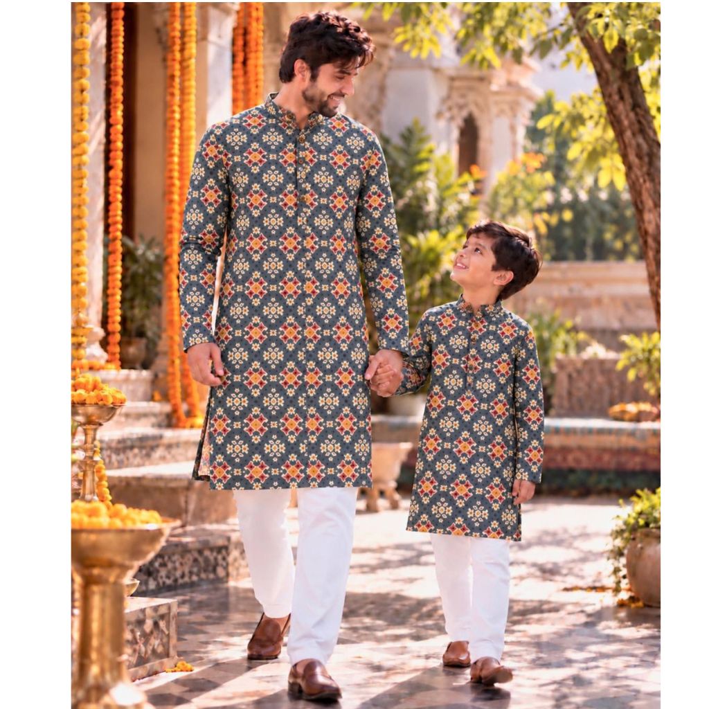 A man and a boy wearing matching printed kurta sets walk hand in hand in an outdoor courtyard. The long-sleeve shirts have a dark teal-blue base with a repeating orange and yellow geometric pattern, paired with white trousers.