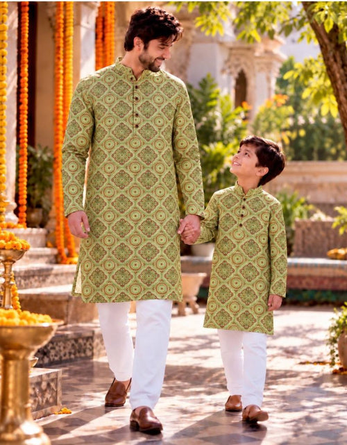 Load image into Gallery viewer, A man and a boy walking hand in hand outdoors, both wearing matching light olive green patterned kurta sets with white trousers. The background shows a sunlit courtyard with marigold garlands, plants, and architectural details.
