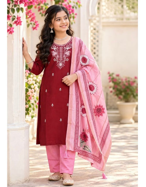 Load image into Gallery viewer, A young girl modeling a traditional Indian salwar kameez suit. The outfit consists of a deep maroon red tunic with white embroidery, pink pants, and a light pink dupatta with red floral patterns, set against a soft-focus outdoor background with white pillars and pink flowers.
