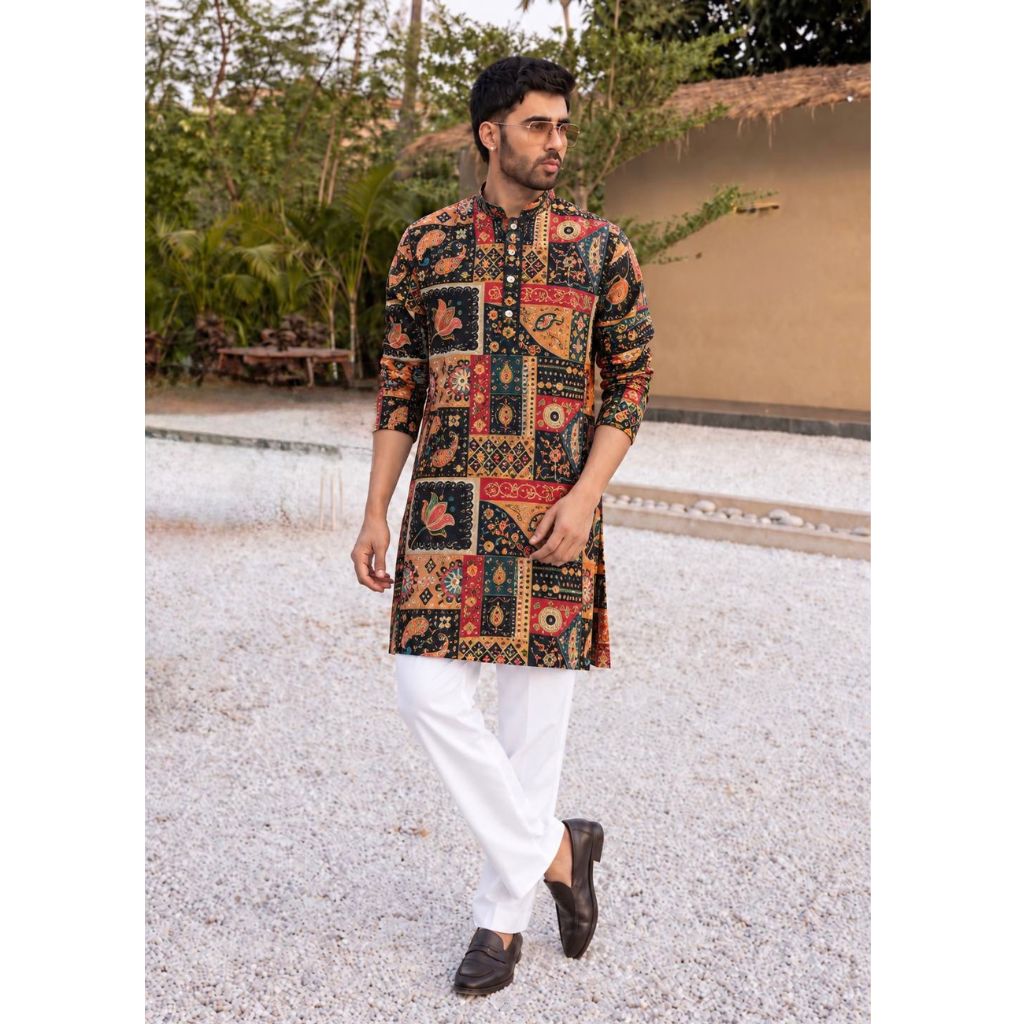 A man wearing a multicolor patchwork kurta with red, orange, and black patterns, paired with white trousers and dark brown loafers, walking outdoors on a gravel path.