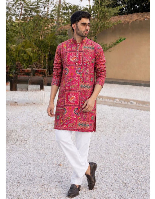 Load image into Gallery viewer, A man wearing a vibrant pink and red patterned kurta with white trousers and dark brown shoes, walking outdoors on a gravel path with trees and a wall in the background.
