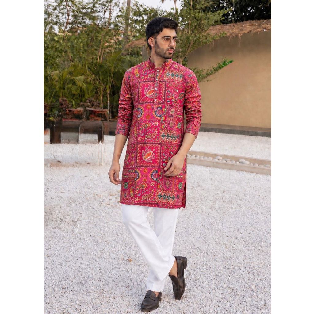 A man wearing a vibrant pink and red patterned kurta with white trousers and dark brown shoes, walking outdoors on a gravel path with trees and a wall in the background.