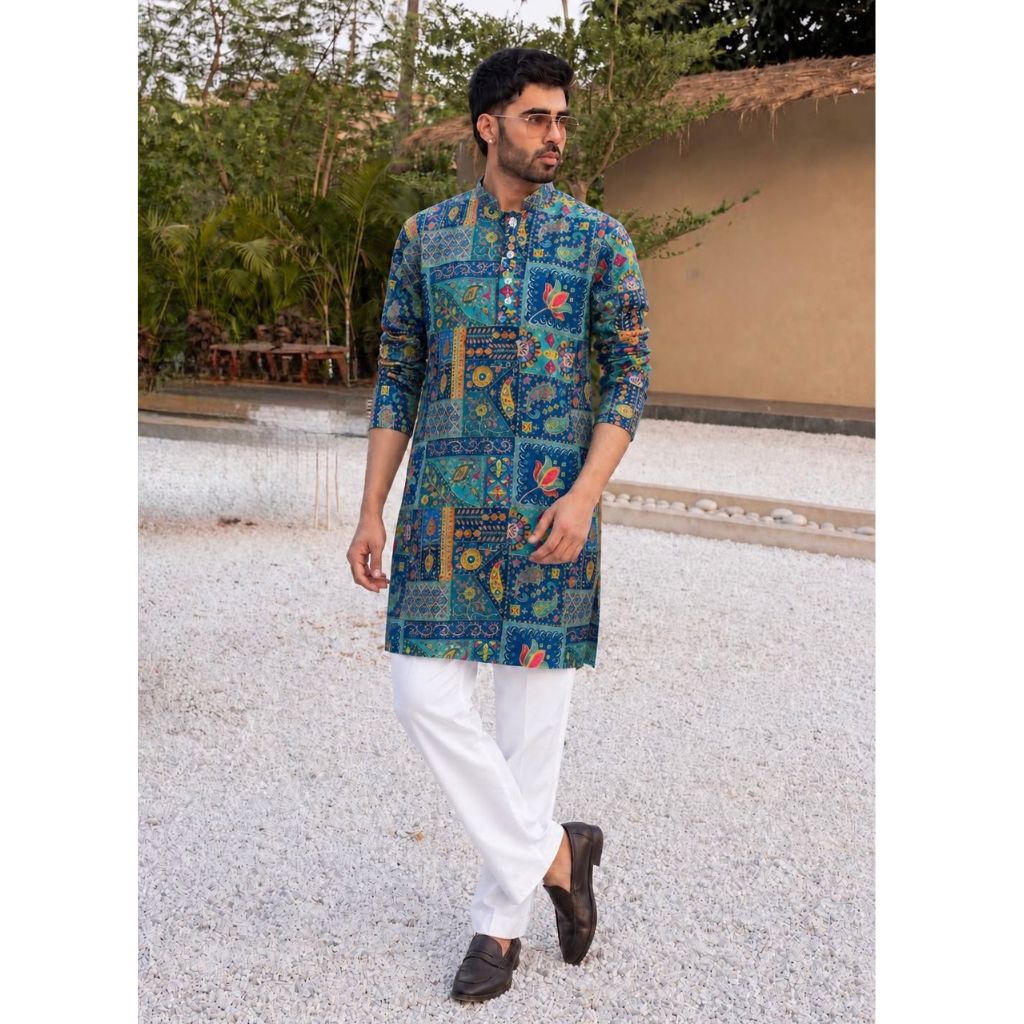 A man wearing a blue and green patterned kurta with white trousers and dark brown shoes, standing outdoors on a light gravel path with a garden and wall in the background.