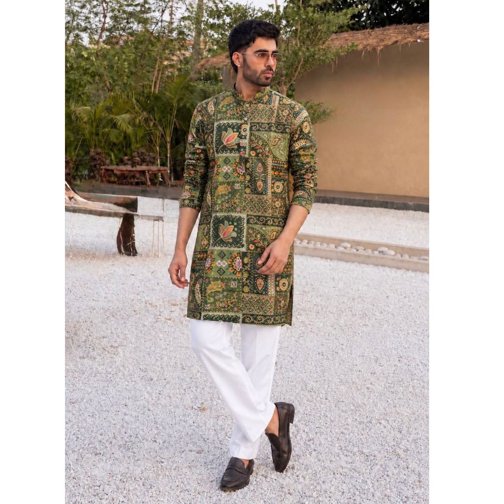 A man wearing a green and gold patterned kurta with white trousers and dark loafers, standing outdoors on a light gravel path.