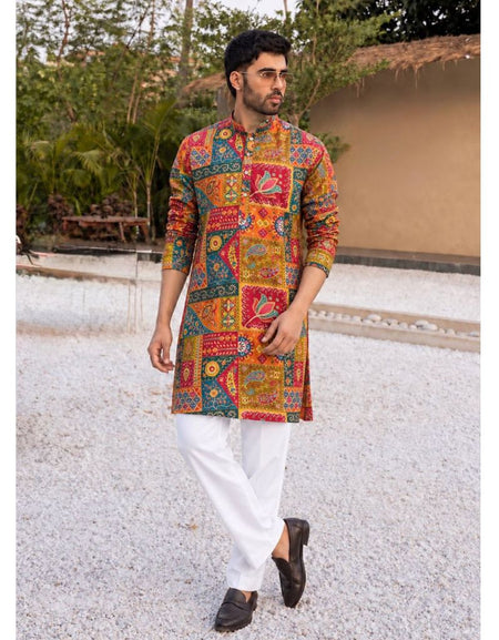 A man wearing a vibrant, multicolored patchwork-print kurta with white trousers and black loafers, standing outdoors on a light gravel path with a house and greenery in the background.