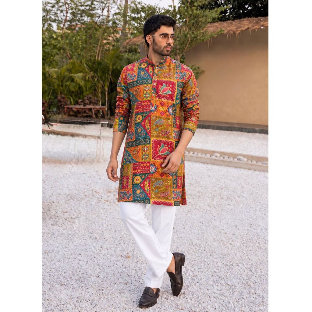 A man wearing a vibrant, multicolored patchwork-print kurta with white trousers and black loafers, standing outdoors on a light gravel path with a house and greenery in the background.