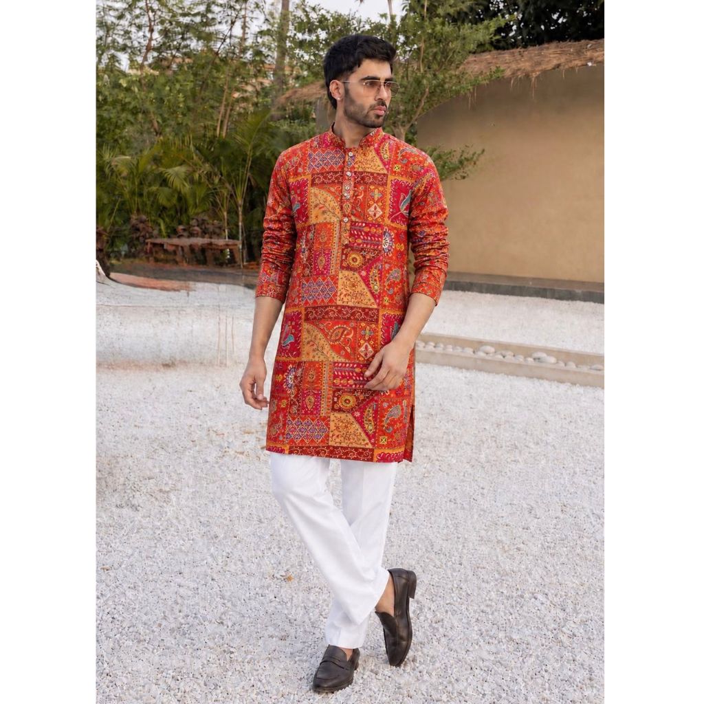 A man wearing a vibrant red and orange patchwork-patterned kurta with white trousers and dark loafers, standing outdoors on a light-colored gravel path.