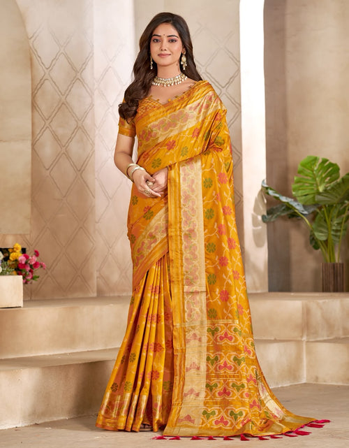 Load image into Gallery viewer, A woman models a golden yellow Banarasi silk saree with intricate red and green floral patterns and a gold zari border, set against a classical stone architectural background.
