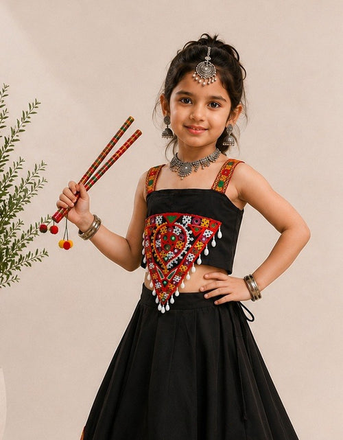 Load image into Gallery viewer, A young girl modeling a black sleeveless traditional dress with colorful embroidered details on the bodice, accessorized with silver jewelry, while holding a pair of red-and-black wooden sticks next to a potted plant against a beige wall.
