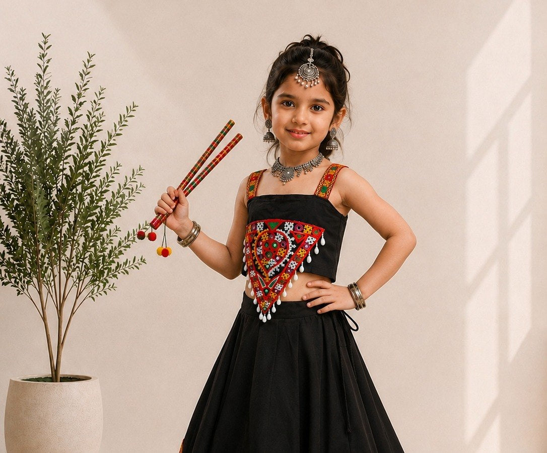 A young girl modeling a black sleeveless traditional dress with colorful embroidered details on the bodice, accessorized with silver jewelry, while holding a pair of red-and-black wooden sticks next to a potted plant against a beige wall.