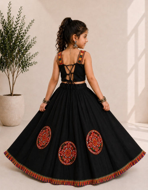 Load image into Gallery viewer, A young girl is shown from the back wearing a black traditional Indian lehenga choli dress with colorful red, orange, and yellow embroidered details. She is standing in a bright, modern room with a potted plant on the left.
