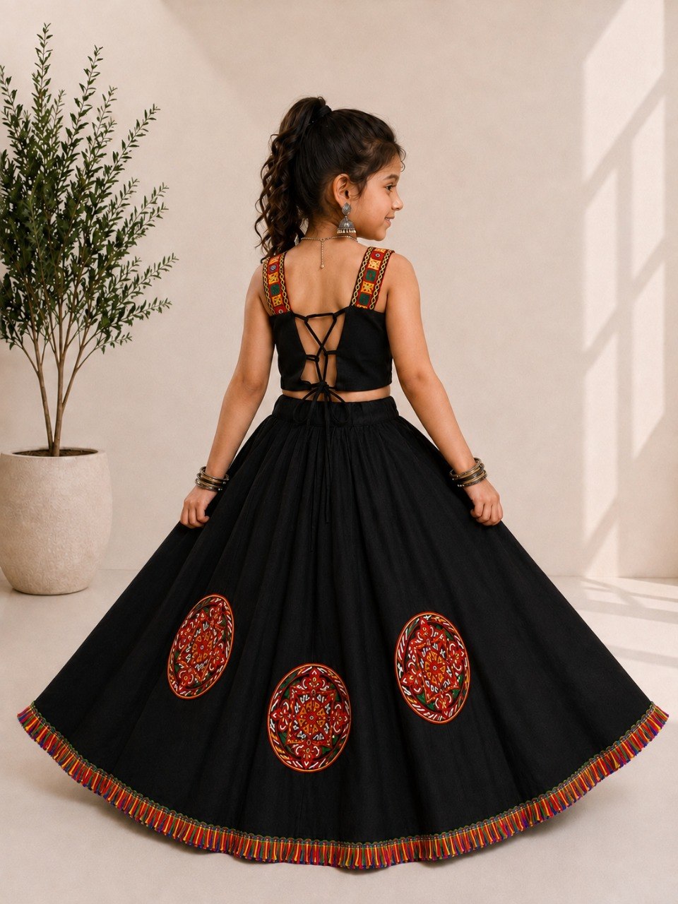 A young girl is shown from the back wearing a black traditional Indian lehenga choli dress with colorful red, orange, and yellow embroidered details. She is standing in a bright, modern room with a potted plant on the left.