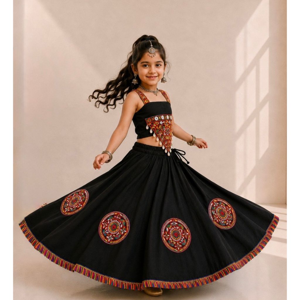 A young girl with long dark hair is wearing a black traditional Indian outfit with ornate red, gold, and silver embroidery, including a sleeveless top and a flared skirt with circular embroidered motifs. She is posing in a studio with a beige background.