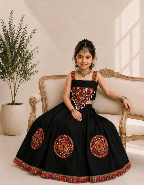 Load image into Gallery viewer, A young girl models a black traditional Indian lehenga choli dress with colorful embroidered details, sitting on a beige sofa in a warm, sunlit room.
