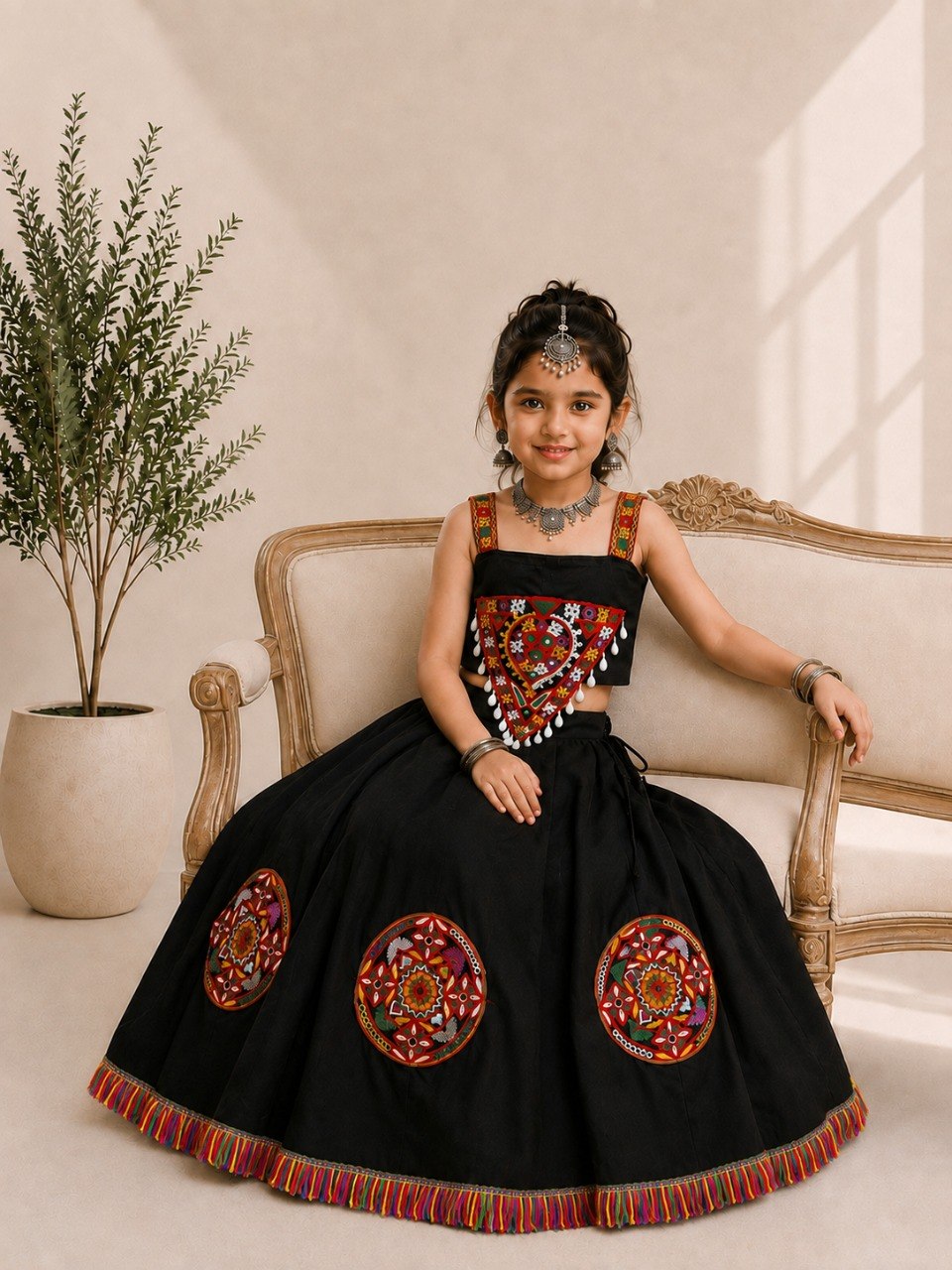 A young girl models a black traditional Indian lehenga choli dress with colorful embroidered details, sitting on a beige sofa in a warm, sunlit room.